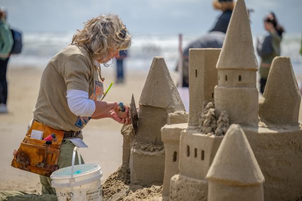 A woman carves details like windows and eves into her sandcastle on the beach.
