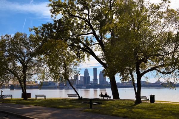 Scenic view of the Louisville skyline across the Ohio River from a waterfront park in Southern Indiana, with large shade trees, park benches and two people sitting by the water.