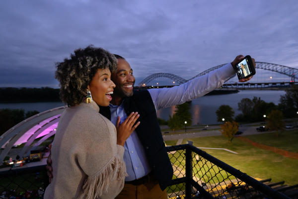 Smiling couple takes a selfie overlooking the Ohio River and Sherman Minton Bridge from the New Albany Amphitheater in New Albany, Indiana, at dusk, with the illuminated amphitheater and waterfront park in the background.