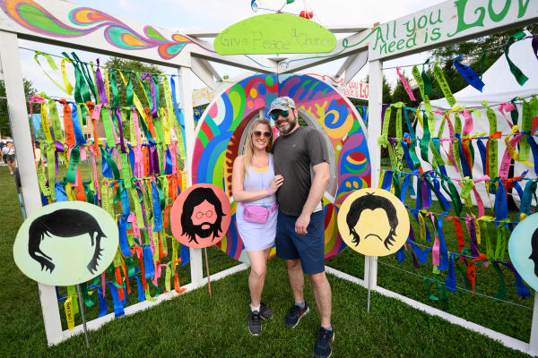 Smiling couple poses in front of a colorful, Beatles-inspired peace and love display decorated with vibrant ribbons, psychedelic artwork and Beatles silhouettes at Abbey Road on the River in Jeffersonville, Indiana.