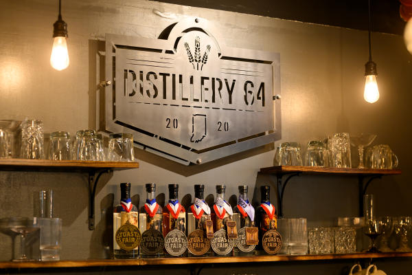 Distillery 64’s metal logo displayed on a wall above a collection of award-winning spirits, medals and glassware, illuminated by warm pendant lighting inside the Southern Indiana distillery.