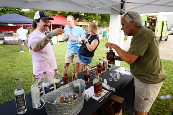 A vendor pours a sample of spirits for an attendee at Distillery 64’s Liquid Legends Fest in Southern Indiana, with bottles of liquor displayed on a tasting table beneath an outdoor tent.