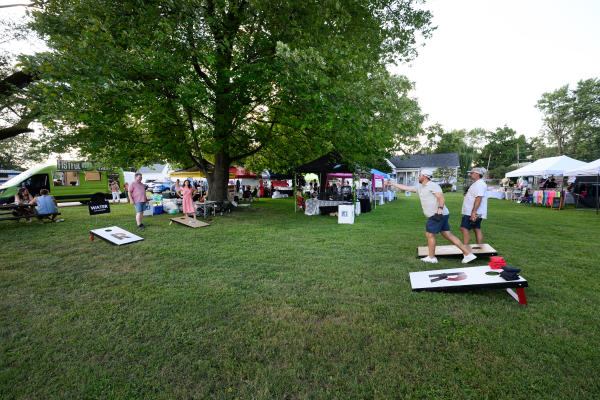 Attendees play cornhole on a grassy lawn at Distillery 64’s Liquid Legends Fest in Southern Indiana, surrounded by vendor tents, a food truck and outdoor seating beneath large shade trees.
