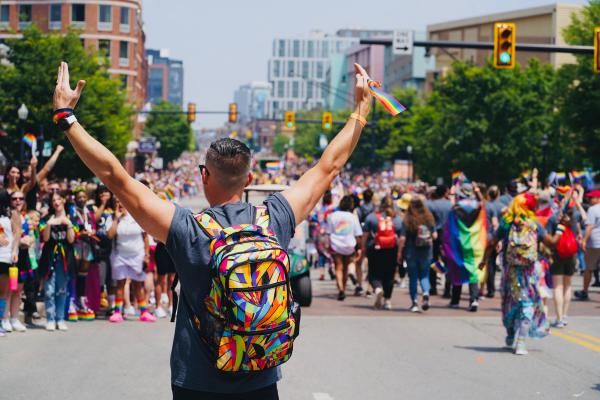 Man with a rainbow backpack has arms in the air while walking in Stonewall Columbus Pride