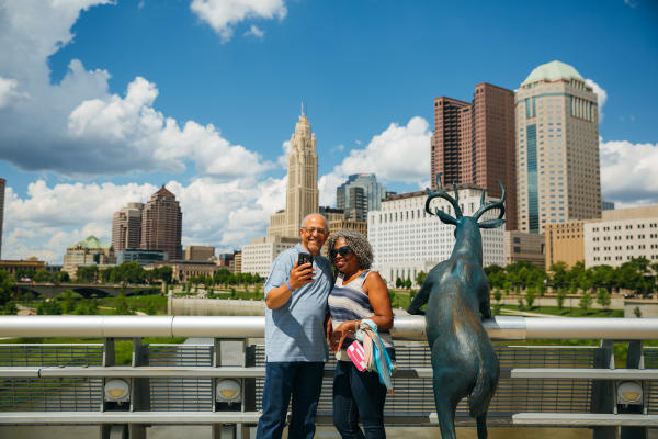 Man and woman taking selfie with deer statue on Scioto River. Columbus skyline in the background.