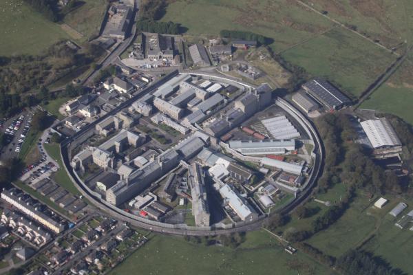Aerial view of a large, circular prison complex with grey buildings and high walls, surrounded by green fields and residential areas, conveying isolation.