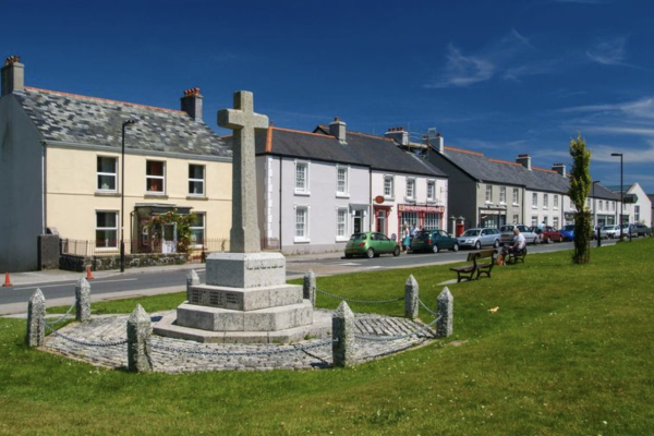 Scenic view of a quaint village with a stone cross monument in the foreground. Behind, coluorful houses line the street under a vibrant blue sky.
