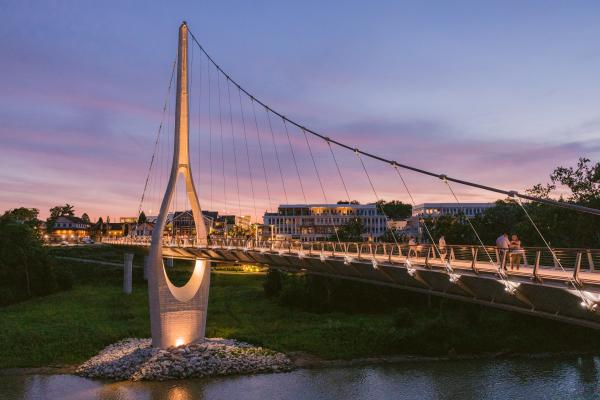The Dublin Link pedestrian bridge illuminated at twilight, with people strolling across and a backdrop of Downtown Dublin, Ohio, under a colorful purple and orange sky.