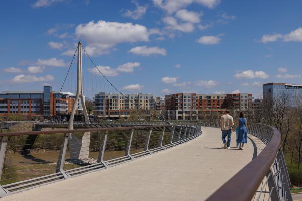 Couple walks hand in hand across Dublin Link bridge with Bridge Park buildings and blue sky in the background.