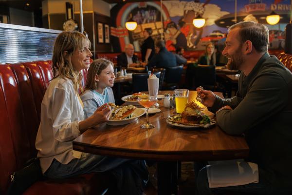 A smiling family enjoys a meal together in a cozy restaurant booth with colorful mural art in the background.