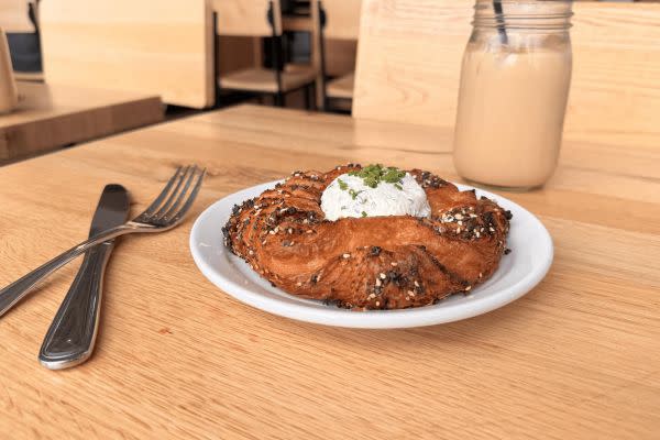 A savory pastry covered in everything bagel seasoning, served with a dollop of whipped cream cheese and garnished with fresh chives on a white plate. The backdrop features the café’s wooden interior and a mason jar with a straw, hinting at a refreshing iced coffee.
