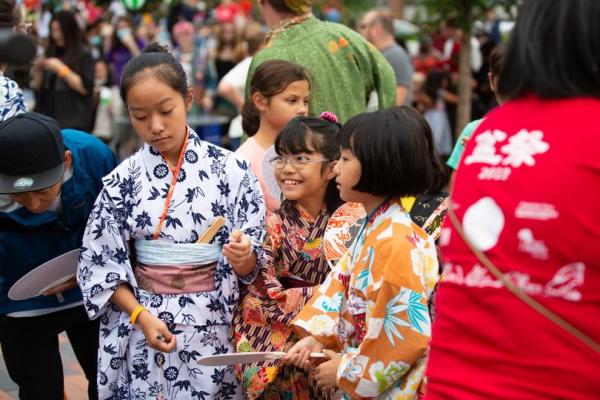 A group of children dressed in colorful traditional Japanese kimonos are participating in an event at Riverside Crossing Park. The children are holding fans and appear to be engaged in an activity together. The background is filled with other event attendees, including a person in a green kimono, all enjoying the lively atmosphere of the park. The event seems to celebrate Japanese culture with various cultural attire and activities.