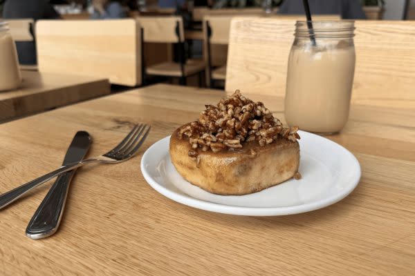 A sweet and sticky cinnamon roll loaded with a generous layer of caramelized pecans, served on a white plate. The setting includes a mason jar of iced coffee and wooden furniture, evoking the warm, inviting atmosphere of the café.
