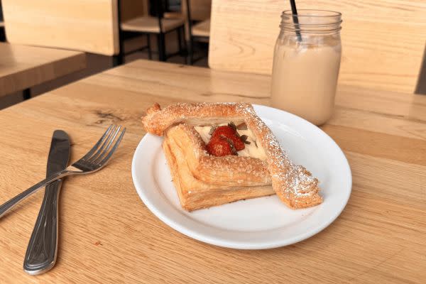 A delicious square pastry topped with powdered sugar, fresh strawberries, and a creamy filling, sitting on a white plate. In the background, there’s a mason jar filled with an iced coffee, adding to the cozy café ambiance with wooden chairs and tables.