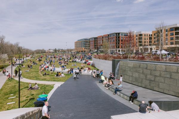 A sunny day at Riverside Crossing Park in Dublin, Ohio, with many people enjoying picnics and relaxing on the grassy area. The park is bustling with activity, with individuals and families scattered throughout. In the background, modern buildings and apartment complexes are visible, creating an urban backdrop for the lively scene. Public restrooms and various amenities are available, and the park is part of the Dublin DORA district.