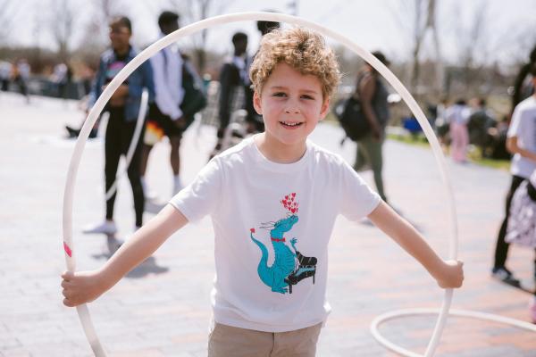 A young boy with curly hair is smiling while holding a hula hoop at Riverside Crossing Park. He is wearing a white t-shirt with a playful graphic of a blue dragon playing a piano and hearts above its head. The background shows other people enjoying activities in the park, with trees and a sunny sky visible in the distance.