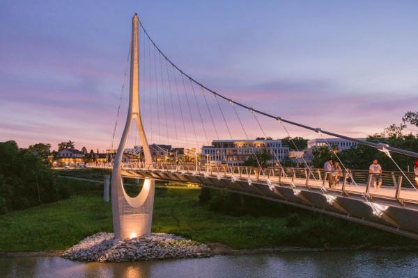 The Dublin Link pedestrian bridge illuminated at sunset, with people walking across and a backdrop of vibrant purple and orange skies over Downtown Dublin, Ohio.