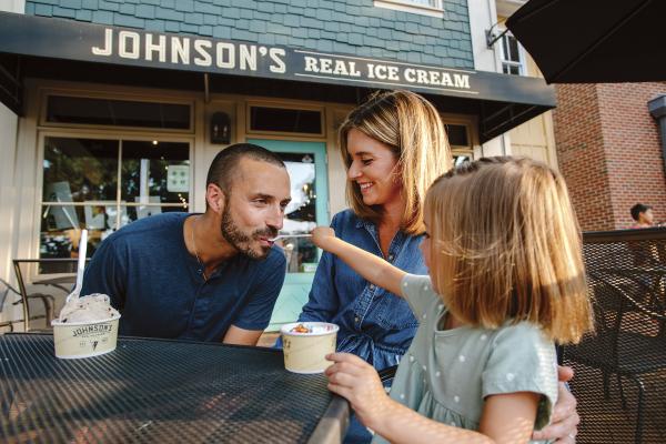 A young daughter feeding ice cream to her dad while her mom watches on the front patio of  Johnson's Ice Cream