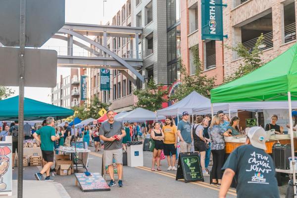 Street market at North Market Bridge Park with vendor tents, people walking and shopping, a man playing cornhole, and signage on nearby buildings. The scene is lively and crowded, with a mix of food and retail booths.