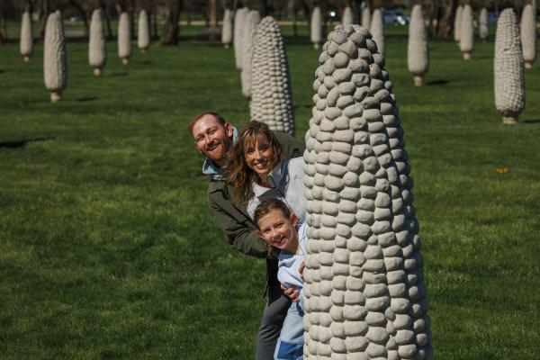 A smiling family peeks out from behind a concrete corn sculpture at the Field of Corn in Dublin, Ohio, on a sunny day.