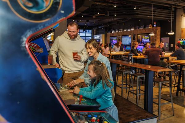 Family playing an arcade game at a bar and arcade in Dublin, Ohio, while enjoying drinks and laughing together.