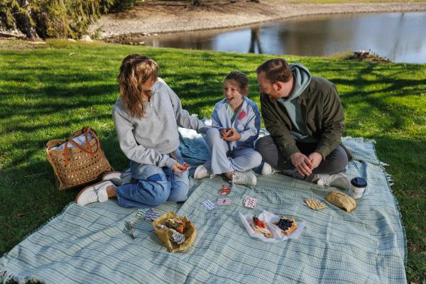 Family enjoys a picnic on a blanket by a pond, laughing and playing cards with food and drinks spread out around them.