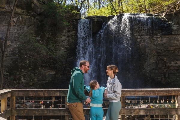 A family smiles and talks together on a wooden viewing platform in front of Hayden Falls in Dublin, Ohio, surrounded by nature.