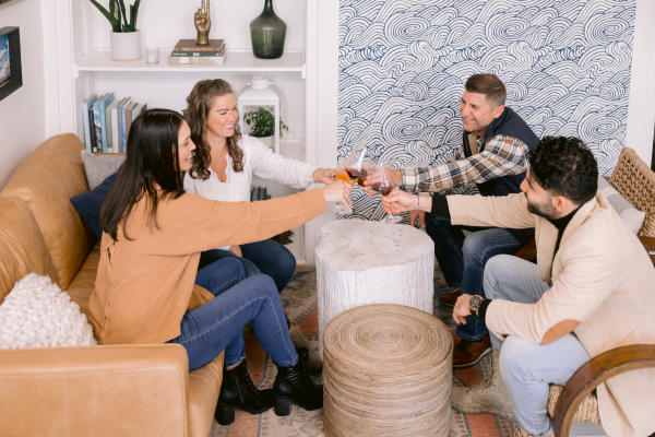 Four friends sitting in Coast Wine house with a leather couch and patterned wallpaper, toasting with glasses of wine around a white tree-stump coffee table. The space features warm tones, bookshelves, and modern decor.