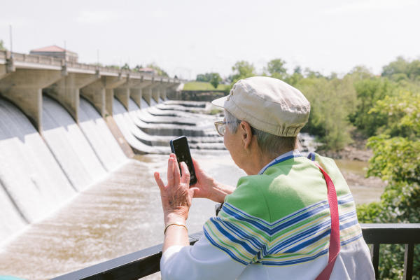 Older woman takes a photo with her phone overlooking a dam with cascading water on a sunny day.