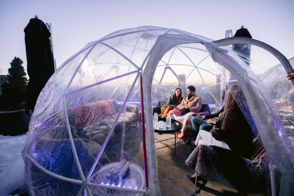 A group of friends relax in a cozy, transparent igloo at VASO Rooftop Lounge with city views.