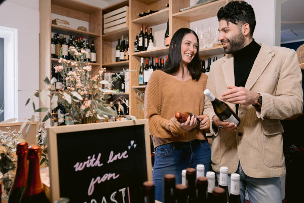 Smiling couple browsing wine bottles in Coast Wine House with wooden shelves filled with various wines and a floral display in the foreground, creating a warm and inviting atmosphere.
