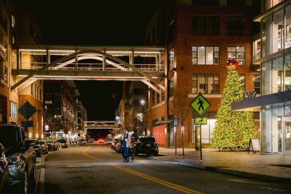 A couple crossing the street at night in Bridge Park next to a giant lit Christmas tree