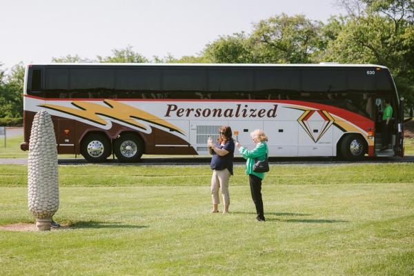 A group tour motorcoach parked at the Field of Corn art installation with visitors taking photos in the field