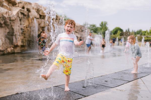 Young boy splashes joyfully through fountains at a splash pad, surrounded by other children playing in the water.