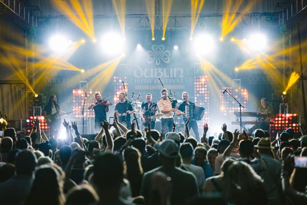 A lively concert at the Dublin Irish Festival featuring the band Skerryvore performing on a brightly lit stage with violins, bagpipes, and an accordion. The crowd is cheering with hands raised in excitement.