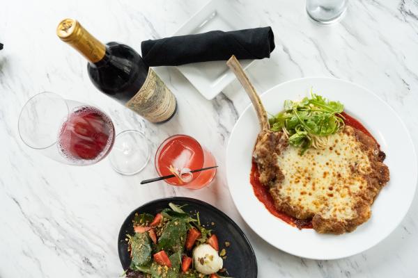 Overhead view of a veal chop with pasta, mixed greens, red wine, and a cocktail at a restaurant with a marble tabletop.