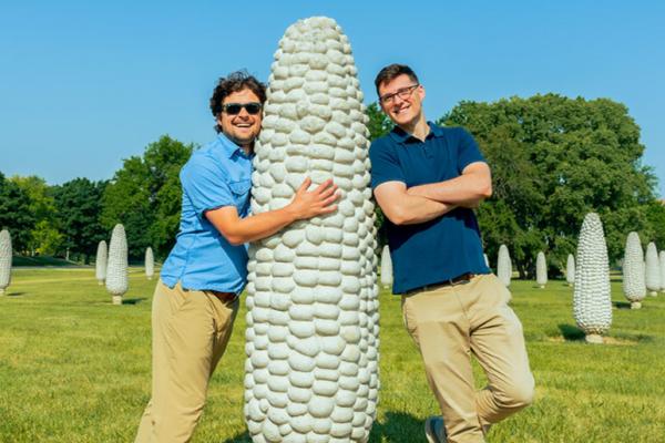 A couple of friends at the Field of Corn. One of them hugging a corn and the other leaning on a corn.
