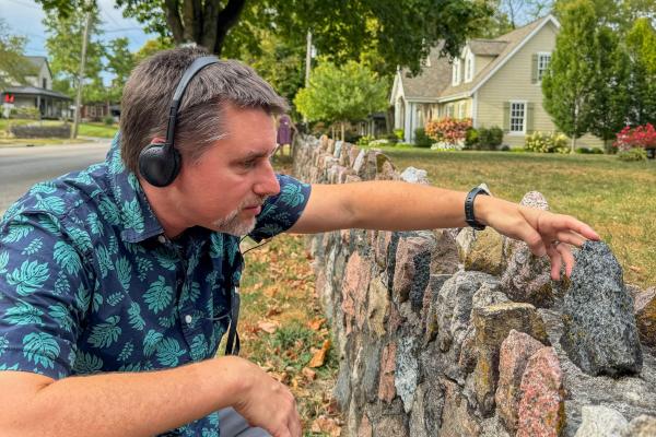 Man wearing headphones and a patterned shirt, examining a stone wall closely with his hand, surrounded by greenery and historic homes in the background. The scene captures a moment of thoughtful observation and engagement with the environment.