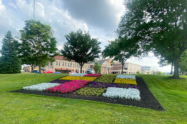 Quilt Garden,Elkhart colorful flower bed arranged in geometric patterns, surrounded by greenery, with buildings and trees in the background under a cloudy sky.
