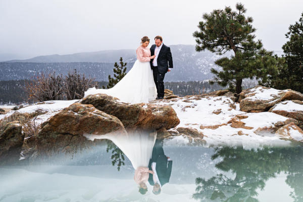A bride and groom share a quiet moment atop a snow-dusted mountain in Estes Park, gazing at their reflection in a still alpine lake. Light snow falls gently around them, capturing the magic of a winter wedding in the Rocky Mountains.