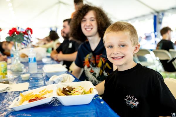 A young boy smiles at the camera while sitting at a long blue-covered table, with a tray of fries and gyro meat in front of him and festivalgoers enjoying food in the background.
