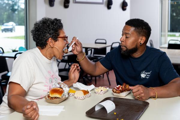 Couple sharing bites at Noble Meats in Cumberland County, NC—man feeds woman with a spoon, with sliders and sides on the table.