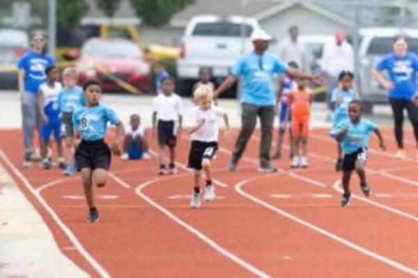 Young children sprint down a red running track during a youth race, with coaches and other participants watching from the sidelines.
