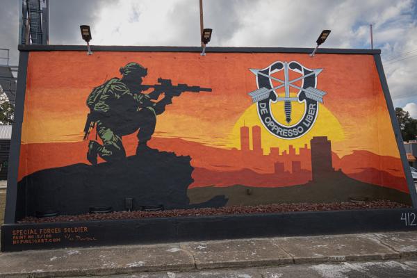A mural of a kneeling soldier in desert camouflage uniform with weapon at the ready, sunset color palette and multiple city skyscapes in the distance