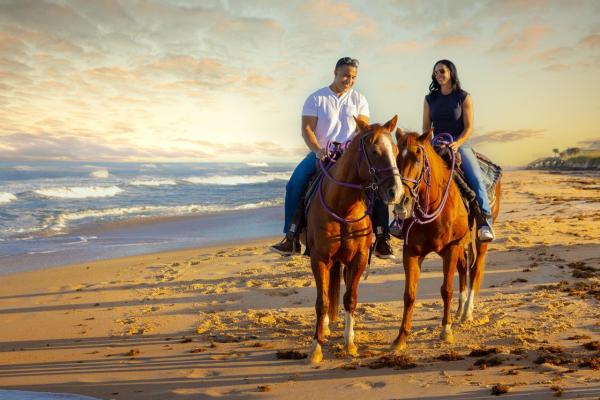 A man and woman ride two brown horses on the beach at Mala Compra Beach Park in Palm Coast and the Flagler Beaches. The man is wearing a white short-sleeve shirt and jeans and the woman is wearing a black sleeveless shirt and jeans. The waves are to the left of them. The horseback ride on the beach is with Equestrian Adventures of Florida in Flagler County, Florida.