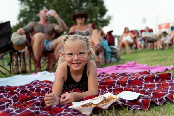 A school-aged girl lays on a blanket in front of her parents, eating chili cheese fries at the Peacemaker Music Fest in Fort Smith.