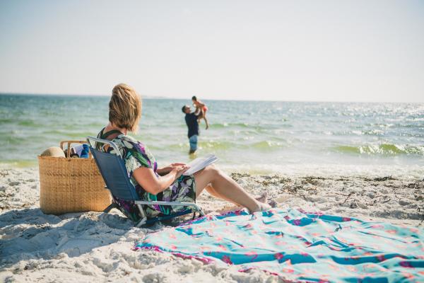 family at the beach. mother sitting with a book on the beach watching dad and daughter play in the water.
