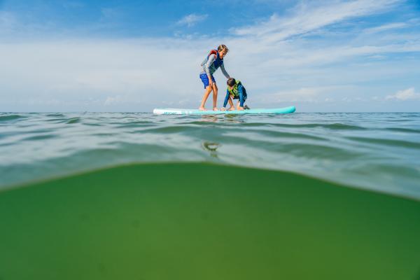 Two children wearing life jackets balance on a paddleboard in calm Gulf County waters under a bright blue sky.