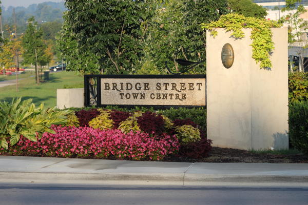 The entrance sign for Bridge Street Town Centre is surrounded by colorful flowers, landscaped greenery, and decorative plantings. The outdoor shopping and entertainment destination is framed by trees and manicured grounds on a sunny day in Huntsville.