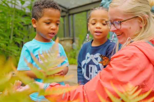 A woman smiles as a monarch butterfly rests on her shoulder inside the Purdy Butterfly House at Huntsville Botanical Garden. Two young children watch with curiosity while surrounded by lush greenery and colorful butterflies.